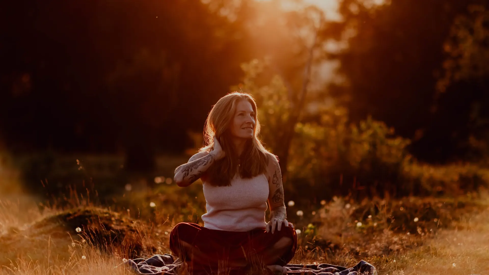 Eve Ragnarsson sitzt im Schneidersitz auf einer Decke in einer weiten Wiese, lächelt und schaut in die tiefstehende Abendsonne.