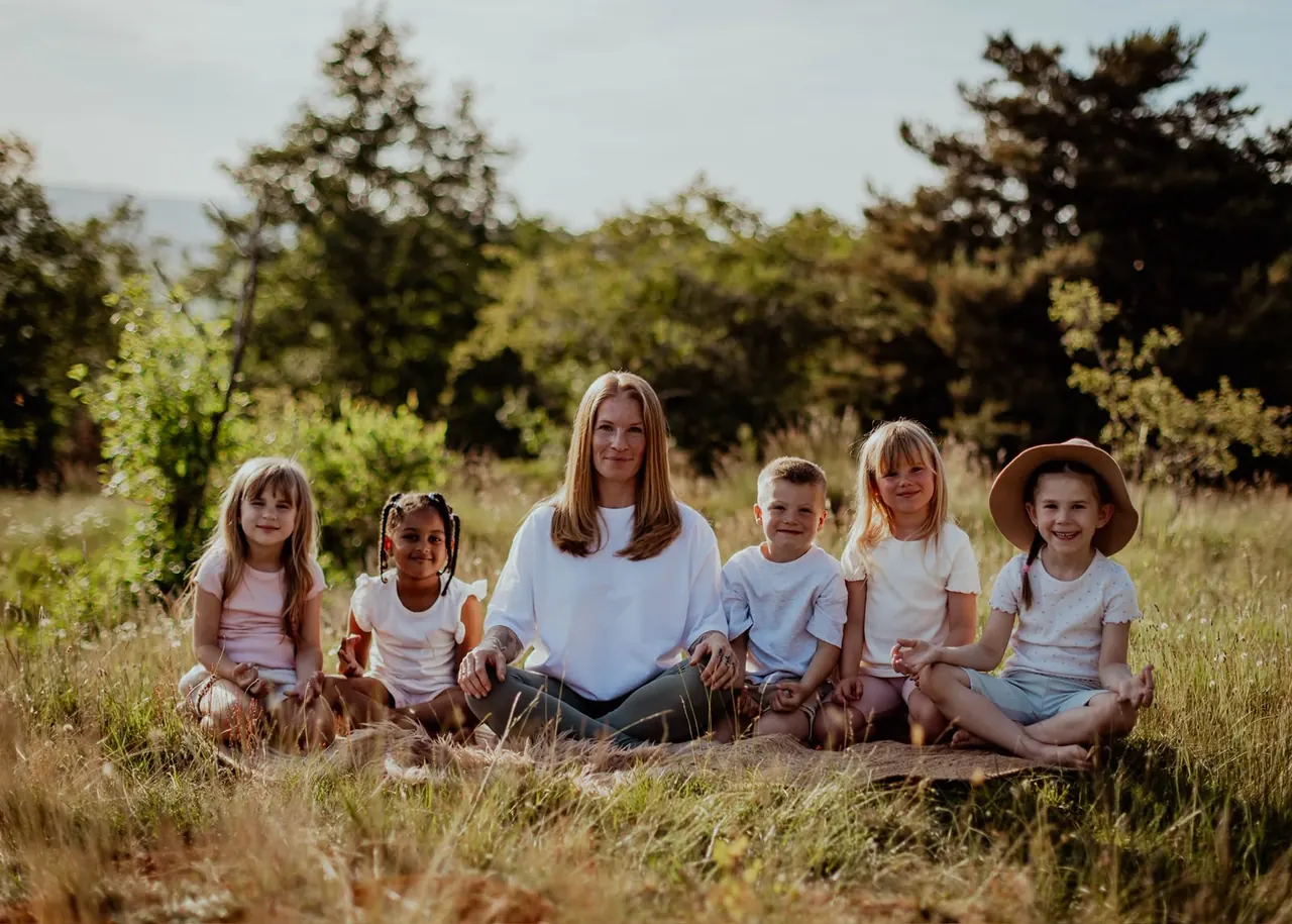 Eve Ragnarsson sitzt im Schneidersitz auf einer Wiese, links und rechts von ihr fünf Kinder in heller Kleidung, alle blicken lächelnd in die Kamera.